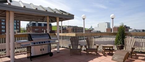 An outdoor patio with a grill, wooden chairs, and tables, overlooking a cityscape.