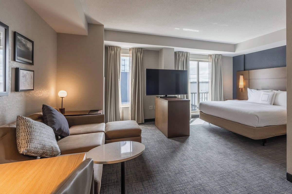 A hotel room with a sofa, a small table, a bed, a TV, and a balcony with curtains.