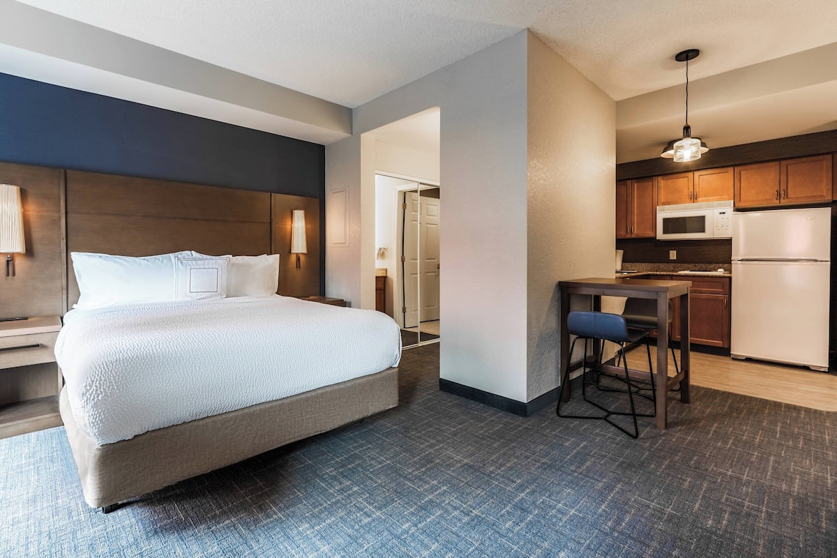 A hotel room with a bed, a desk, a chair, a kitchenette, and a bathroom.