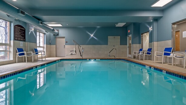 Una piscina con agua cristalina, rodeada de sillas blancas con cojines azules y un cartel que indica las normas de la piscina.