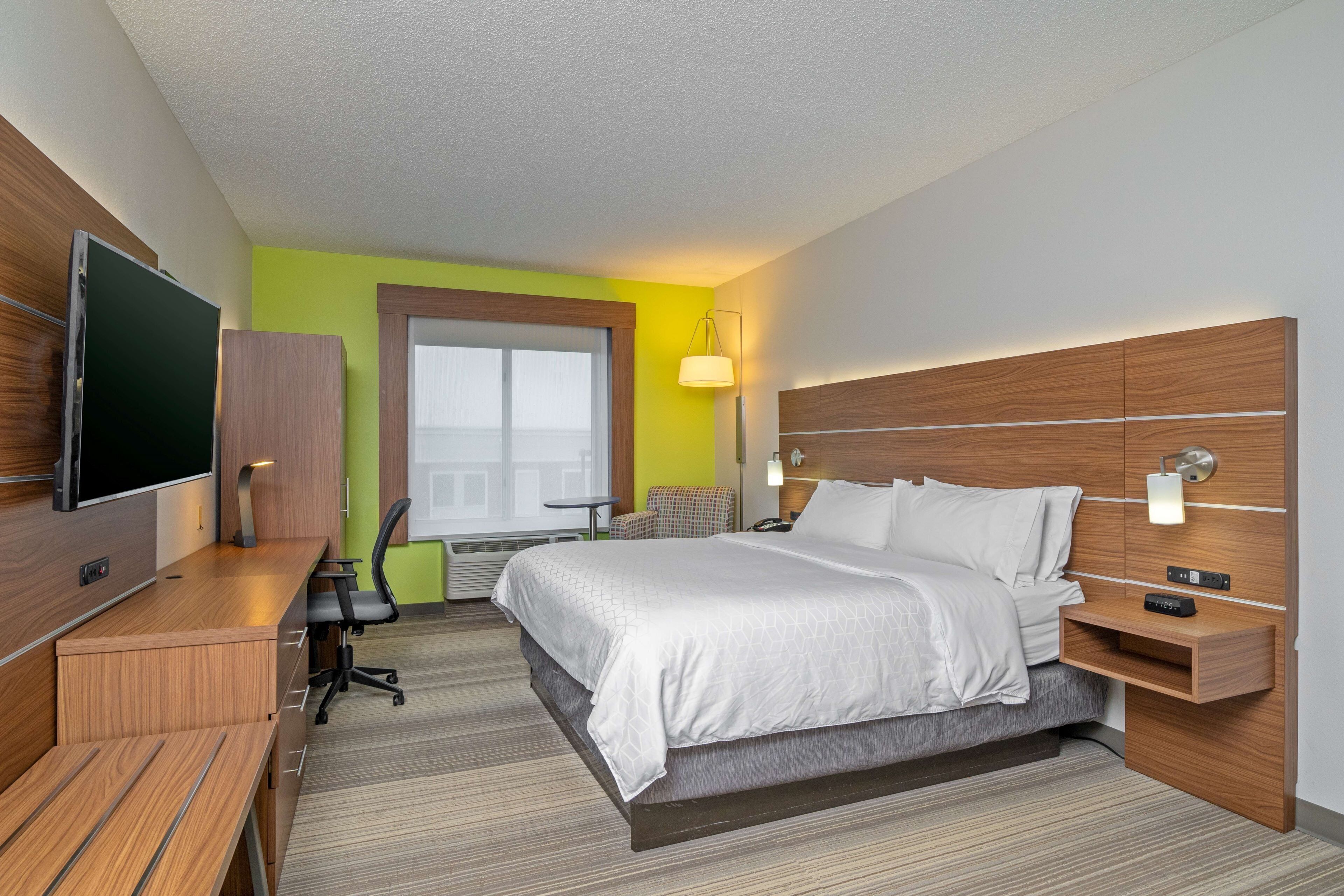 A modern hotel room with a large bed, a desk with a flat-screen TV, a chair, and a bedside table with a lamp.