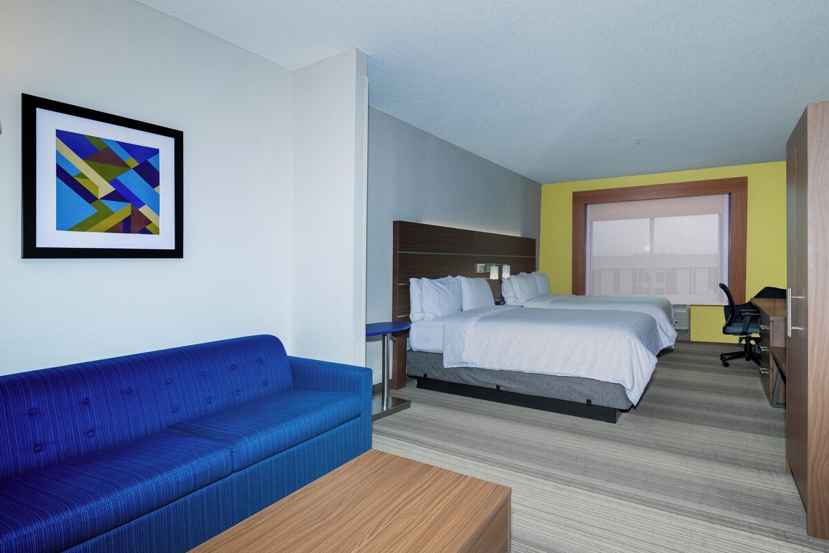 A hotel room with a blue sofa, a bed with white bedding, a wooden desk, and a framed abstract painting on the wall.