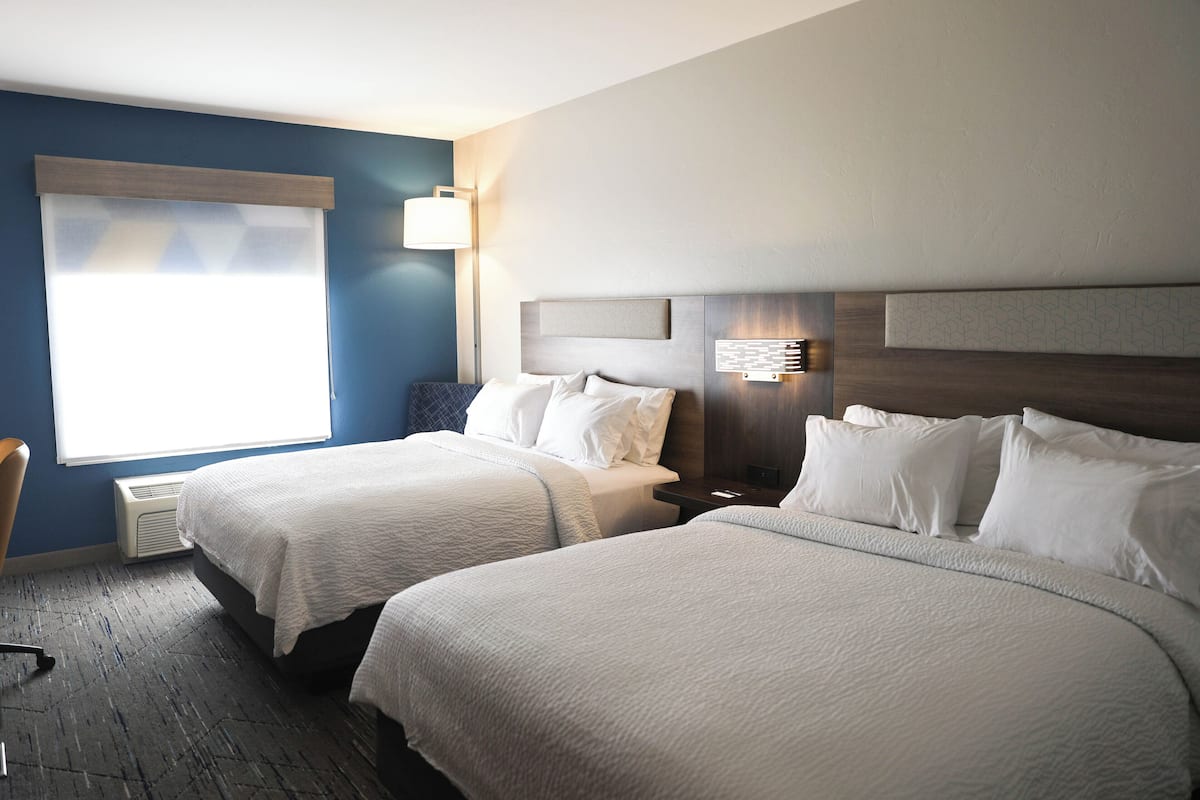 Habitación de hotel con dos camas, un escritorio, una silla y un ventanal grande con persianas.