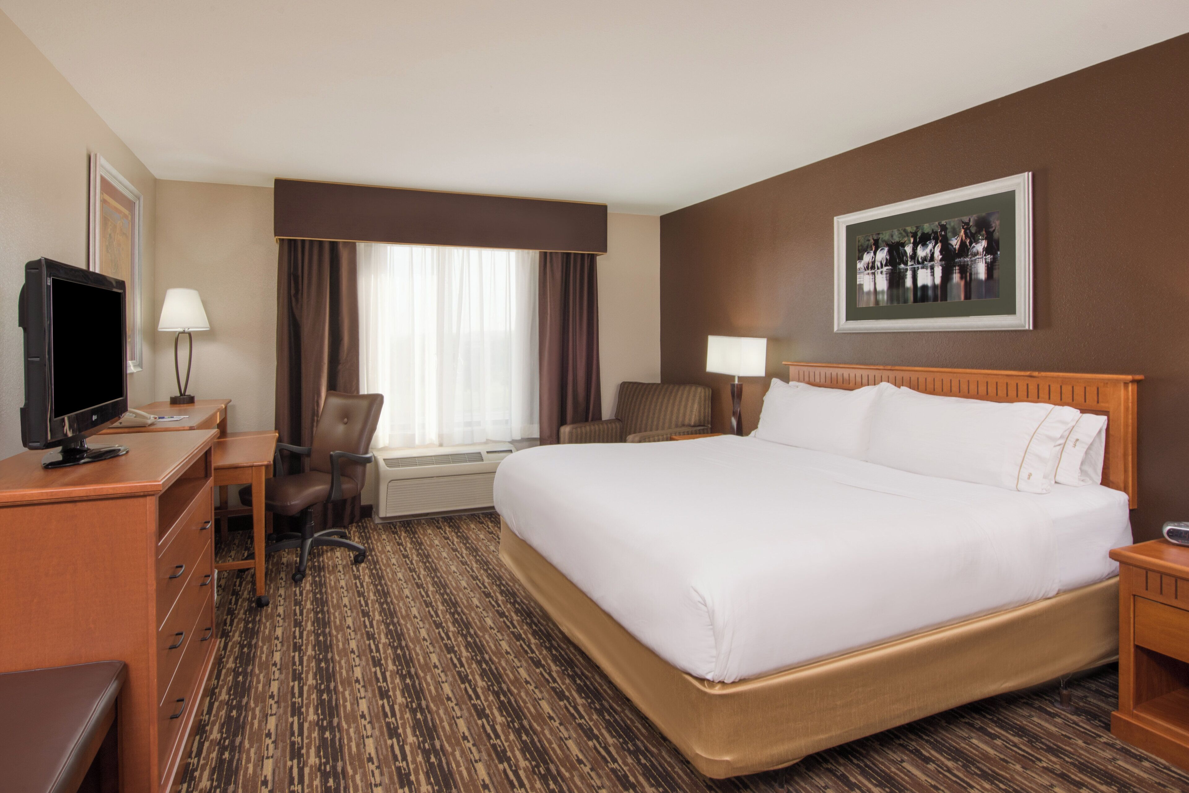 A hotel room with a large bed, a desk with a computer, a chair, a television, and a window with curtains.