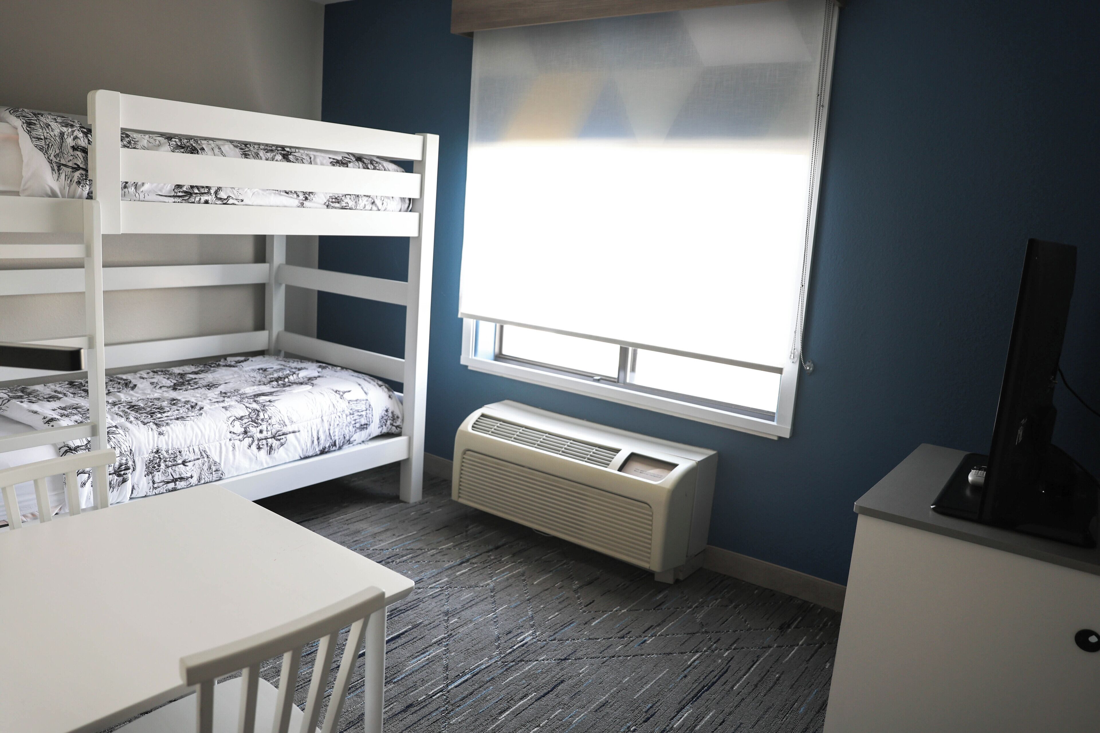 A bunk bed with a patterned bedspread, a desk with a chair, a radiator, and a window with blinds.