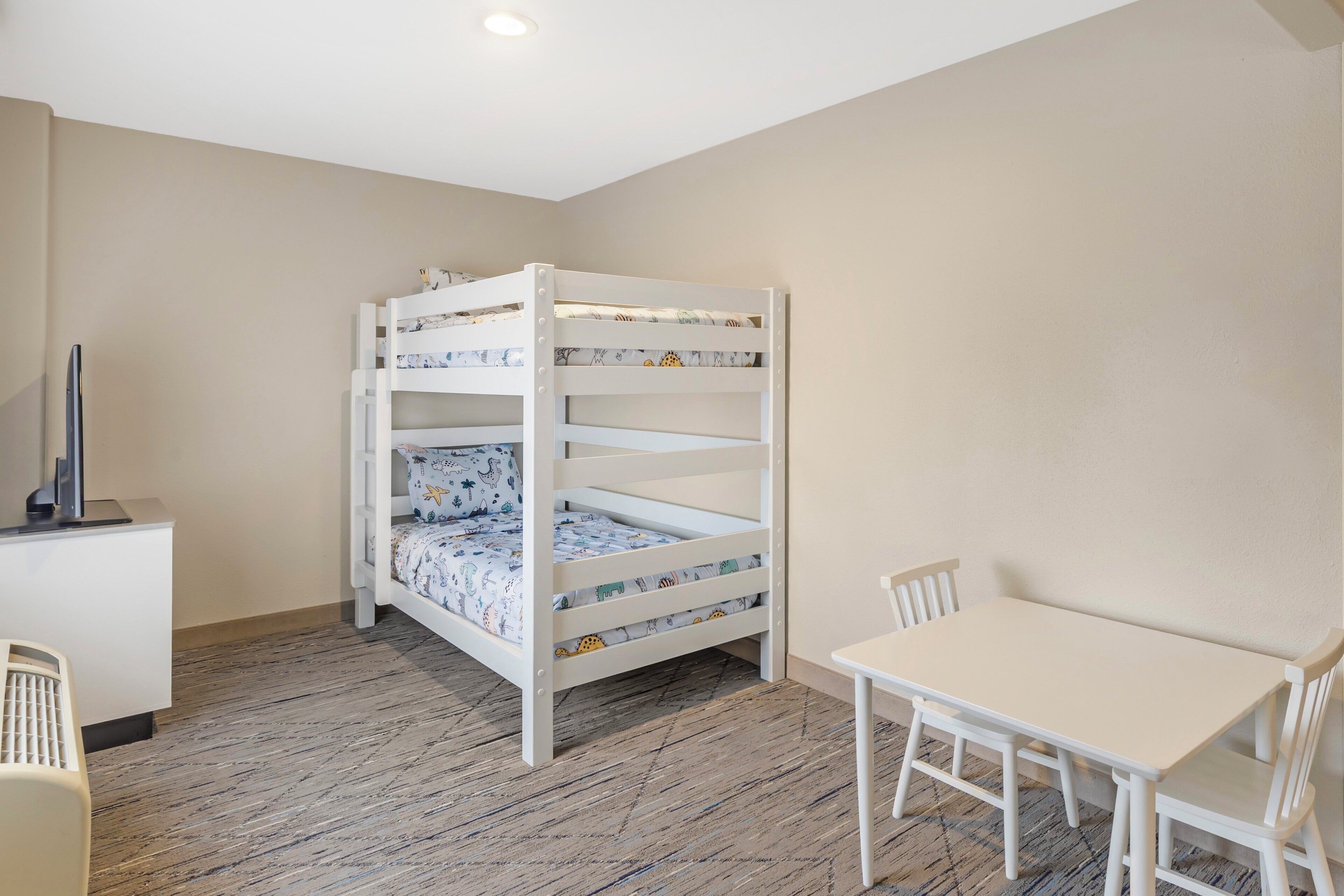 A bunk bed with patterned bedding, a desk with a computer, and a white table with chairs in a room with wooden flooring.