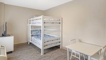 A bunk bed with patterned bedding, a desk with a computer, and a white table with chairs in a room with wooden flooring.