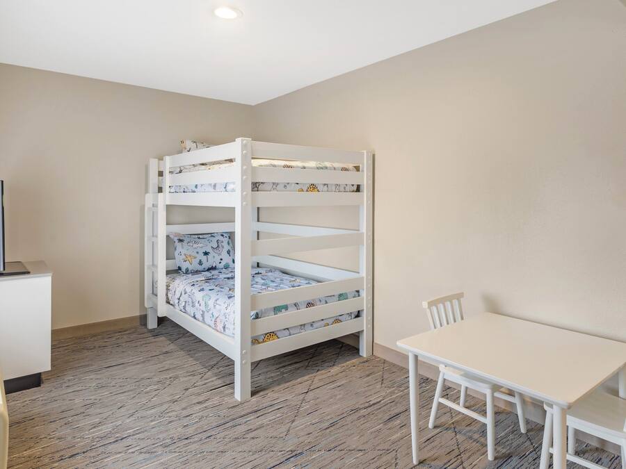 A bunk bed with patterned bedding, a desk with a computer, and a white table with chairs in a room with wooden flooring.