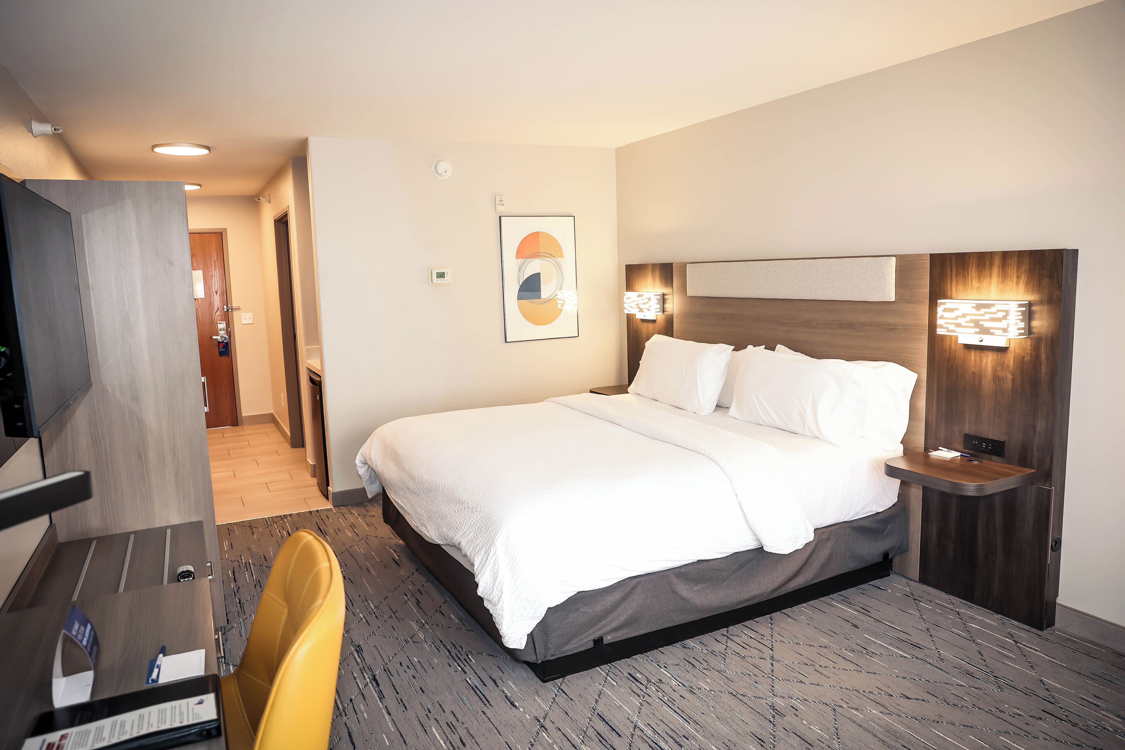 Una habitación de hotel con cama, mesita de noche, silla y escritorio con computadora.