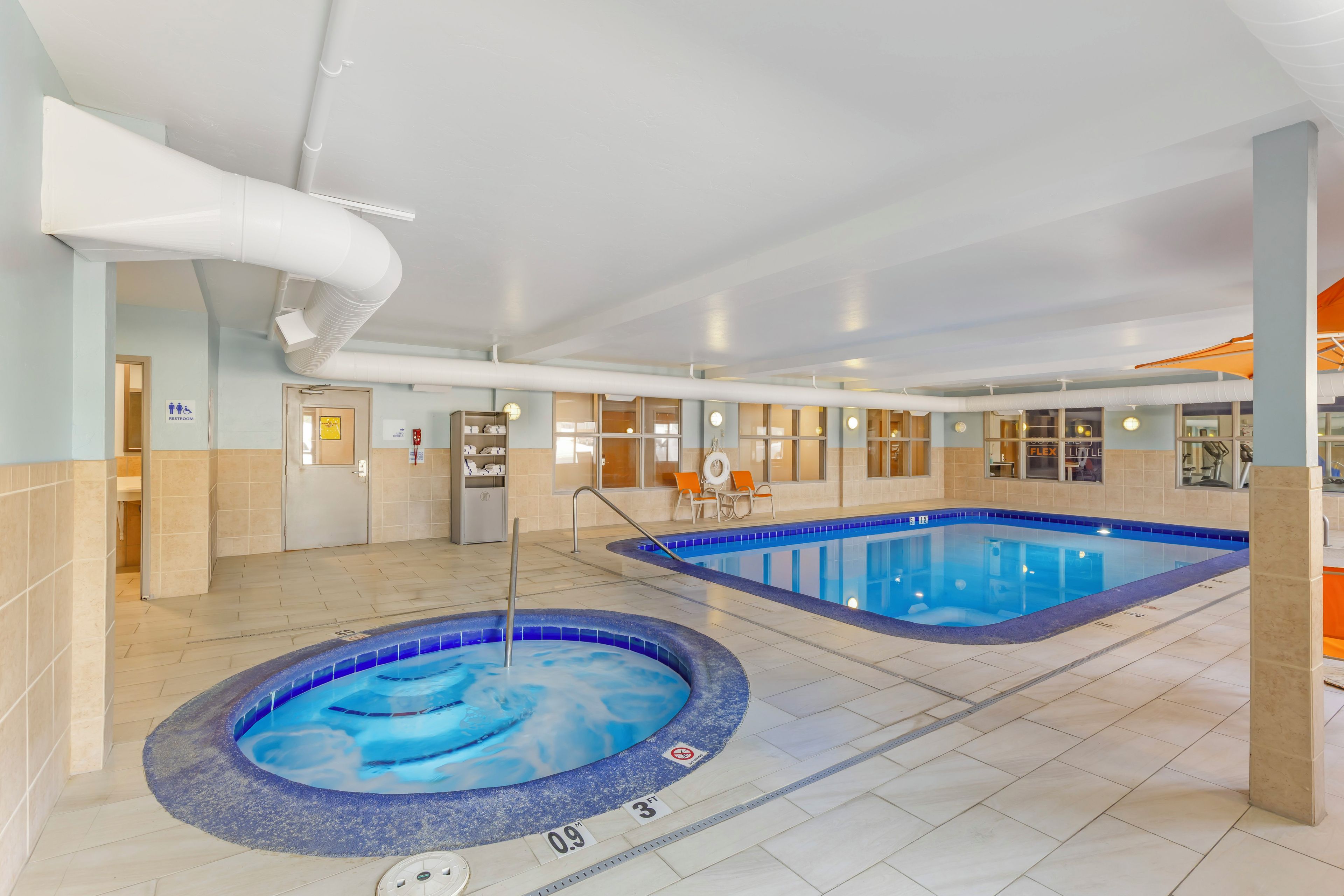 An indoor swimming pool area with two pools, a changing room, and a seating area with a view of the pool.