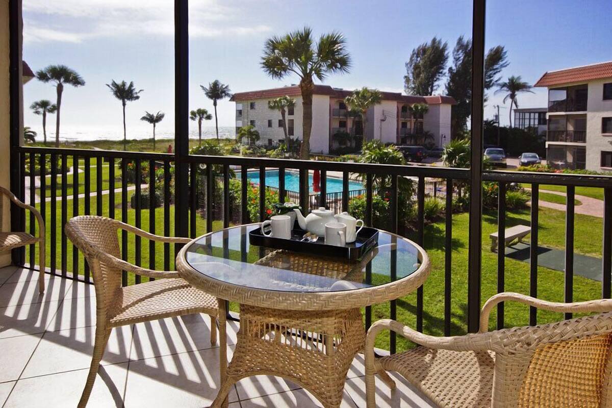 A balcony with wicker chairs and a glass table, overlooking a pool and buildings.