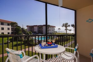 A balcony with a table set for two, overlooking a swimming pool and a building with palm trees.