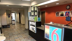 A reception area with a counter displaying the 'motel 6' logo, framed certificates on the wall, and a clock above the counter. - Motel 6 Avoca, IA (Avoca)