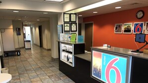 A reception area with a counter displaying the number 6, framed certificates on the wall, and a clock above the counter. - Motel 6 Avoca, IA (Avoca)