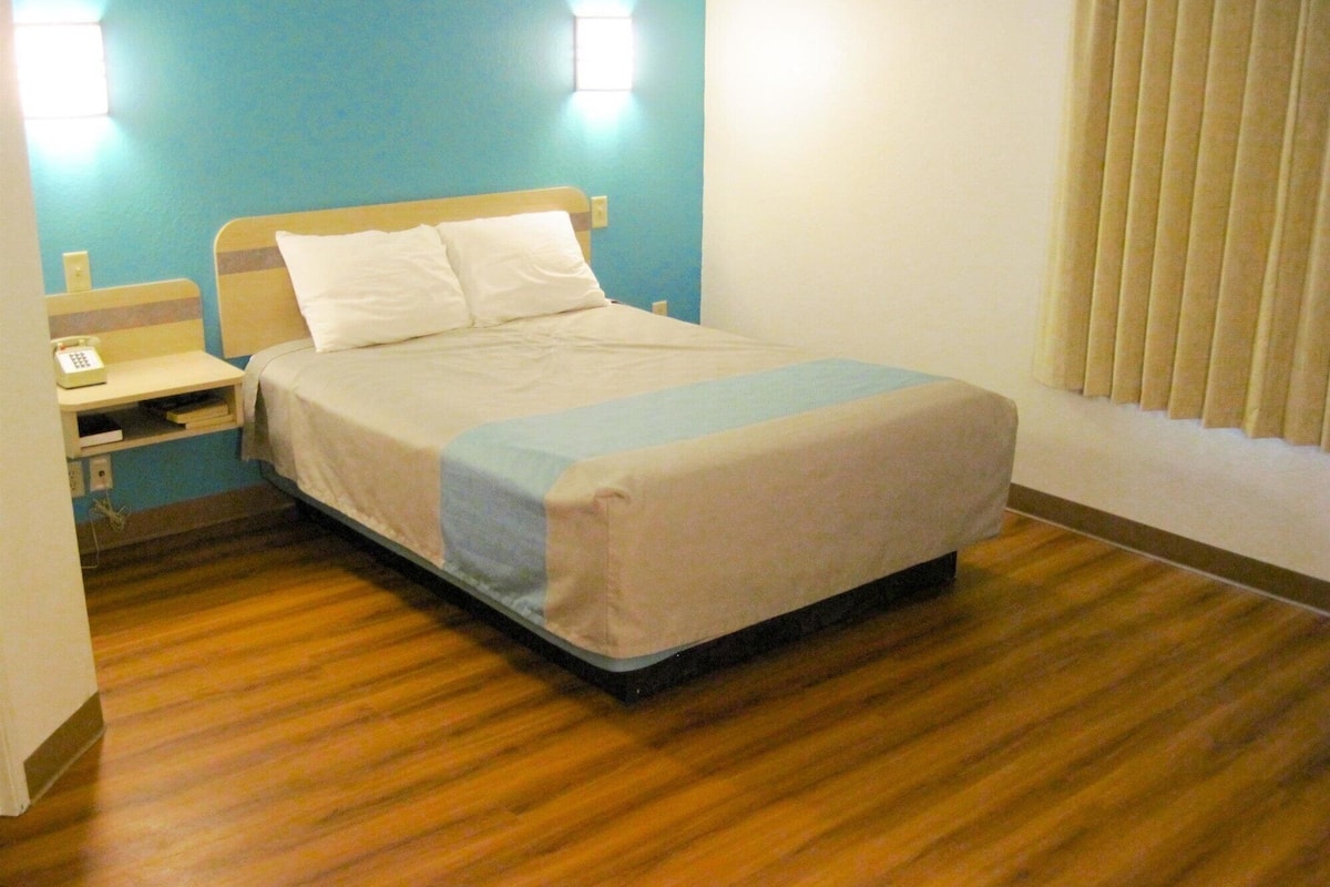 A hotel room with a bed, a nightstand, a lamp, and a window with blinds.