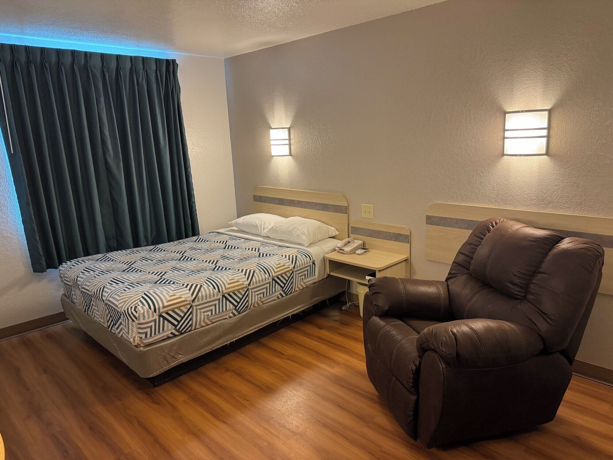 A hotel room with a bed, a nightstand, a brown leather chair, and two wall-mounted lights.