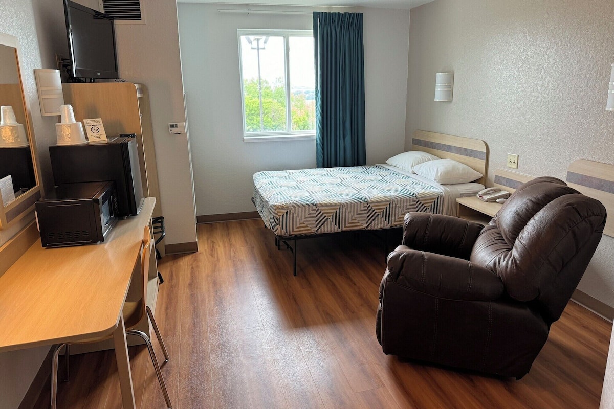 A hotel room with a bed, a desk with a computer, a chair, and a TV on a stand.