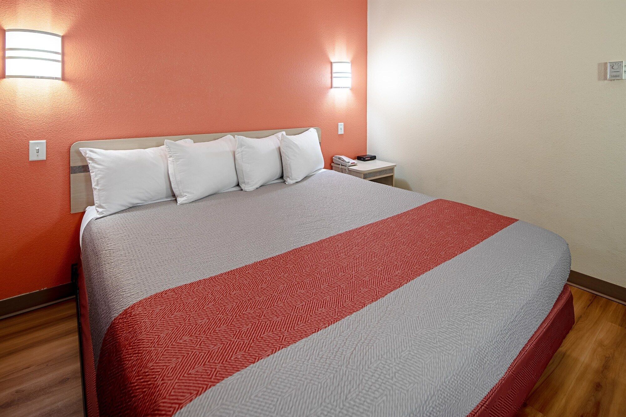 A neatly made bed with a grey and red patterned bedspread, a wooden headboard, and a nightstand with a phone and a book.