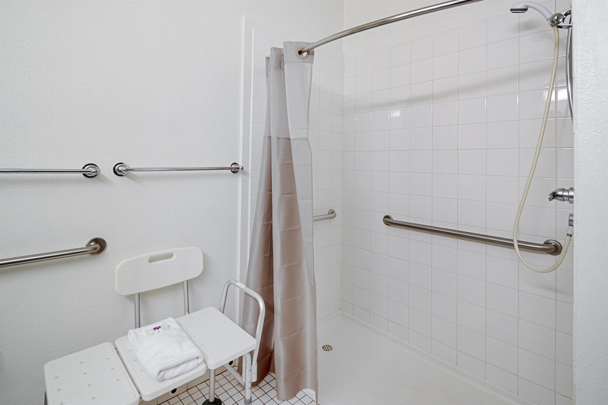 Un baño con azulejos blancos, una ducha con puerta de cristal, un asidero blanco y una toalla blanca sobre un taburete pequeño.