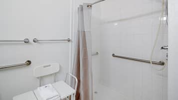 Un baño con azulejos blancos, una ducha con puerta de cristal, un asidero blanco y una toalla blanca sobre un taburete pequeño.