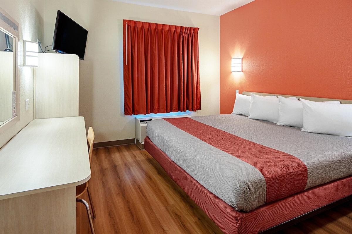 A hotel room with a bed, a desk, a chair, a TV, and a window with red curtains.