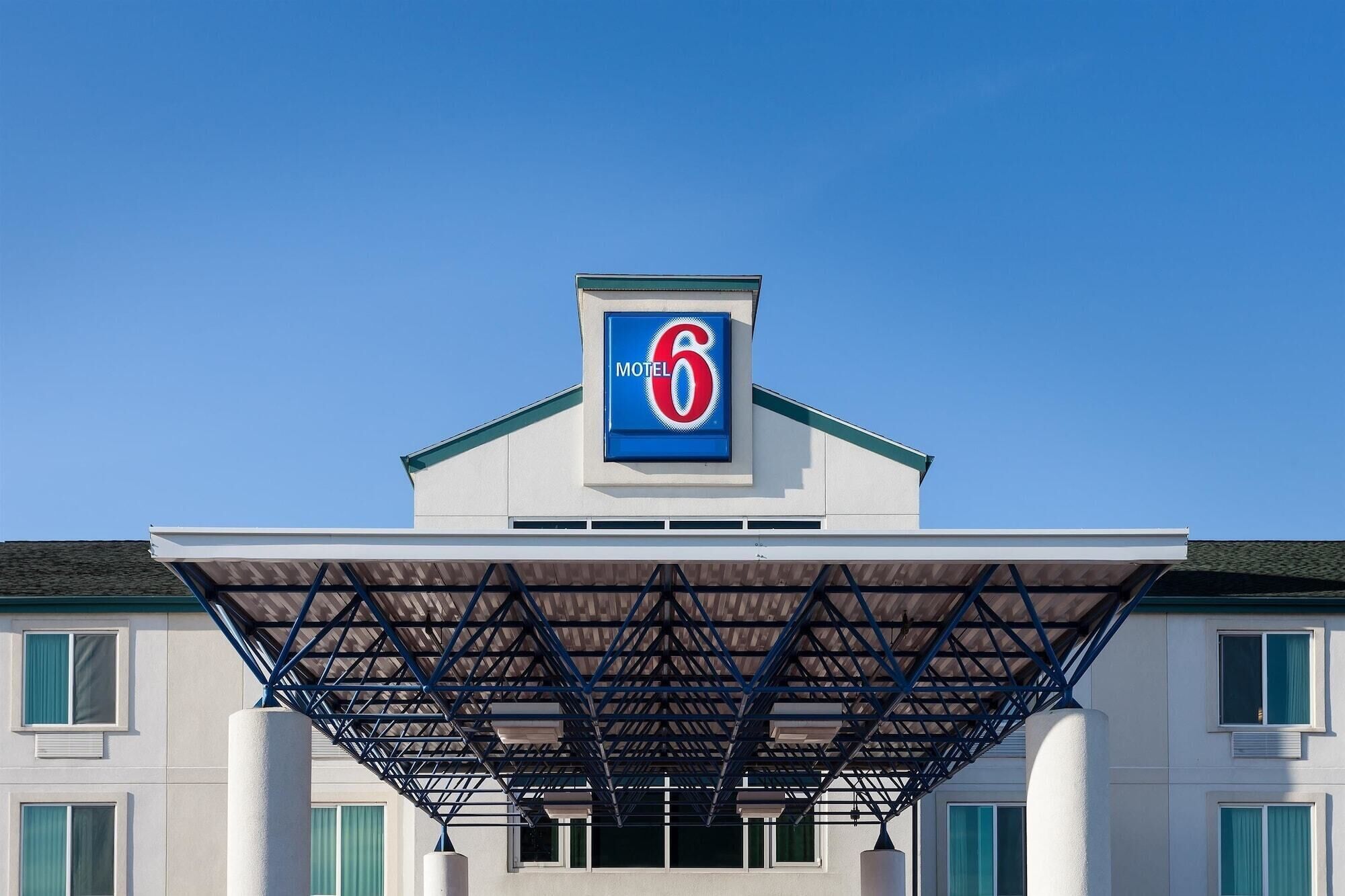 Un motel reconnaissable à son grand panneau « 6 » sur le toit, à son entrée couverte dotée d’une structure métallique et à sa façade blanche ornée de cadres de fenêtres verts.