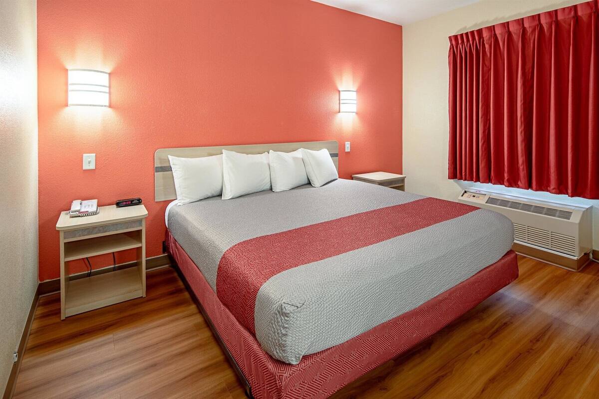 A hotel room with a bed, nightstands, a heater, and a window with red curtains.