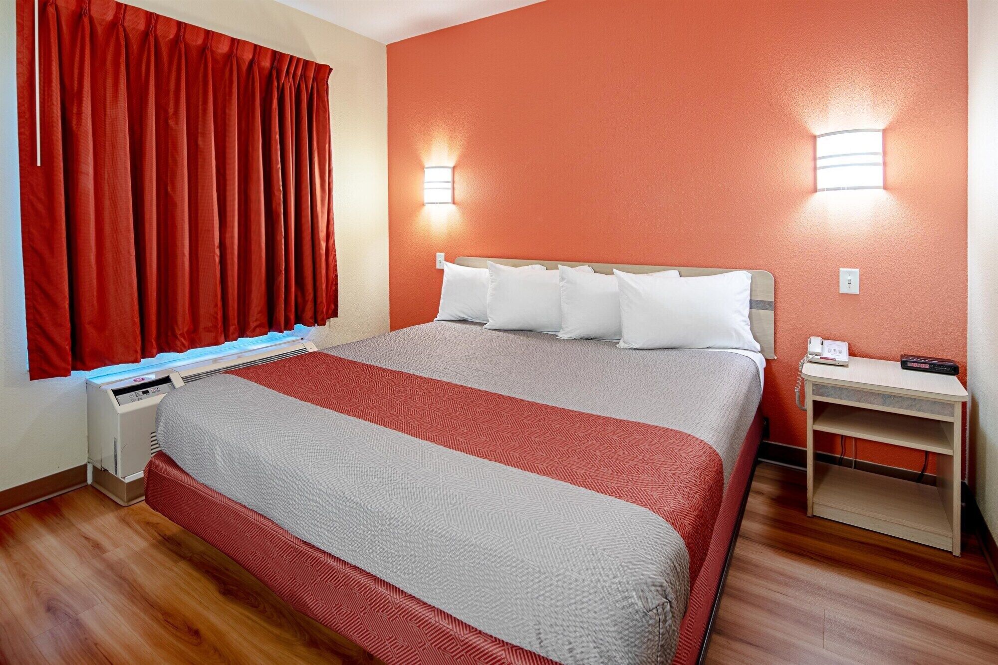 A hotel room with a bed, red curtains, a heater, a bedside table with a phone, and a wall-mounted light.