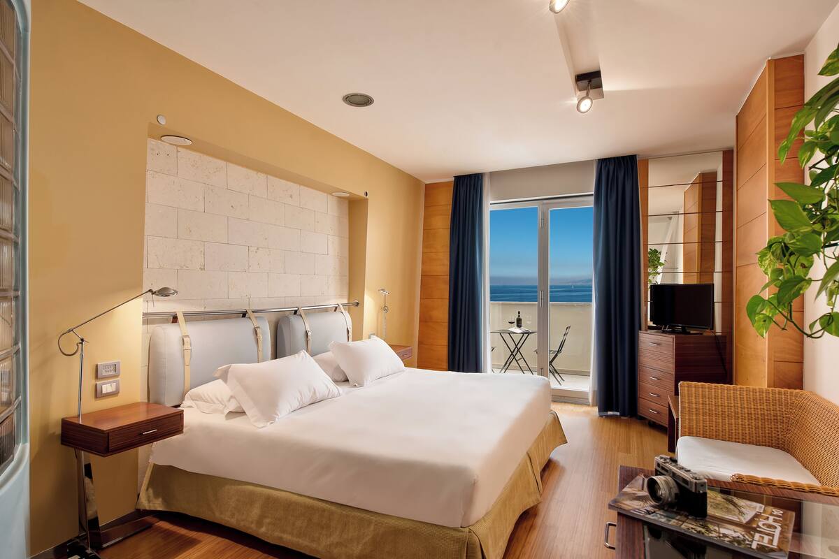 Ein Hotelzimmer mit einem großen Bett, einem Fernseher, einem kleinen Tischchen mit einer Kamera und einem Balkon mit Meerblick.
