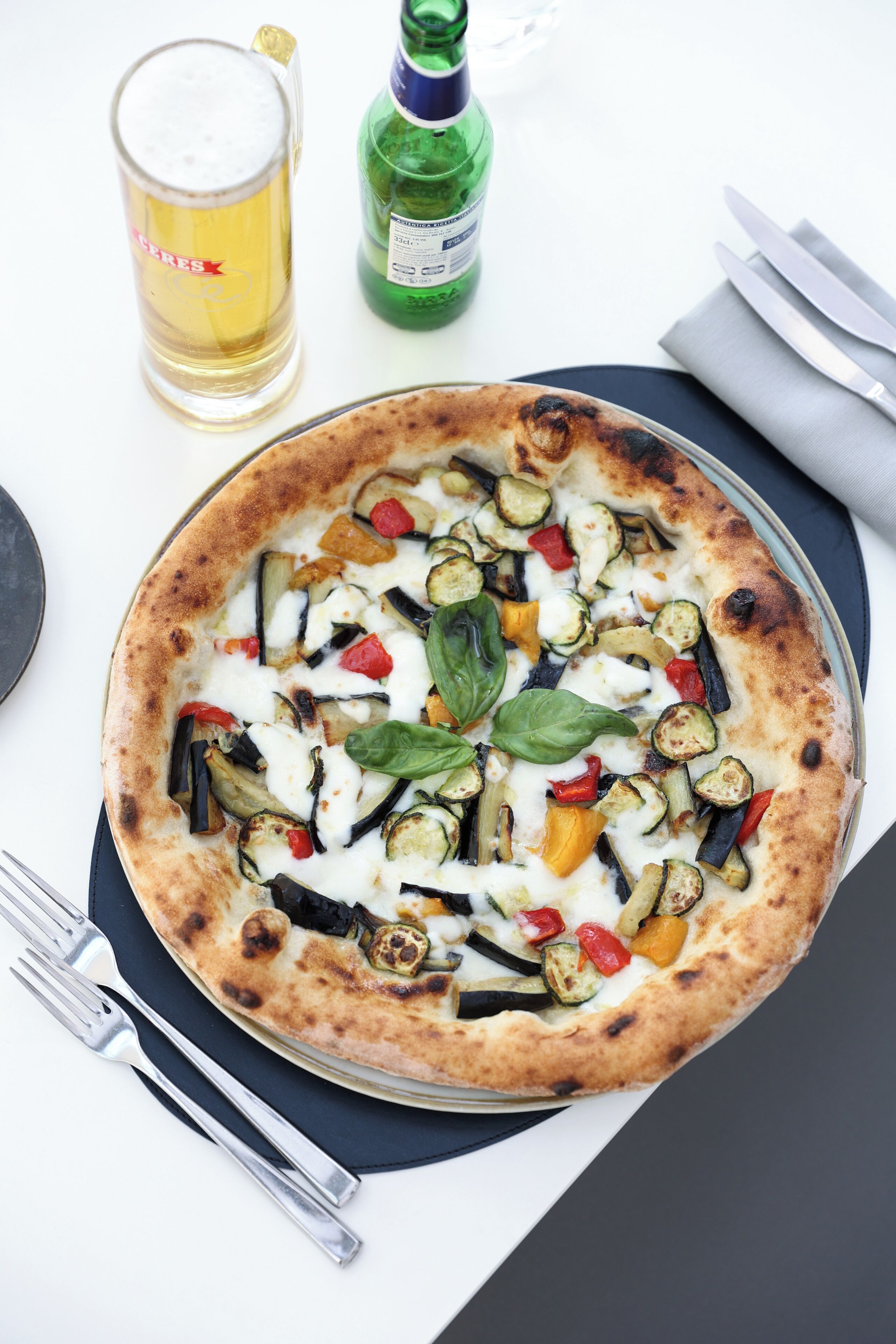 A pizza with various vegetables on a plate, accompanied by a glass of beer and a bottle of water.