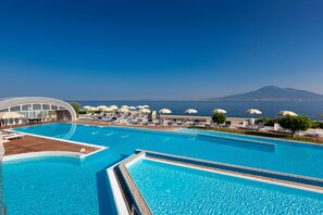 A large outdoor swimming pool with lounge chairs and umbrellas, a wooden deck, and a view of the sea and a mountain in the background.