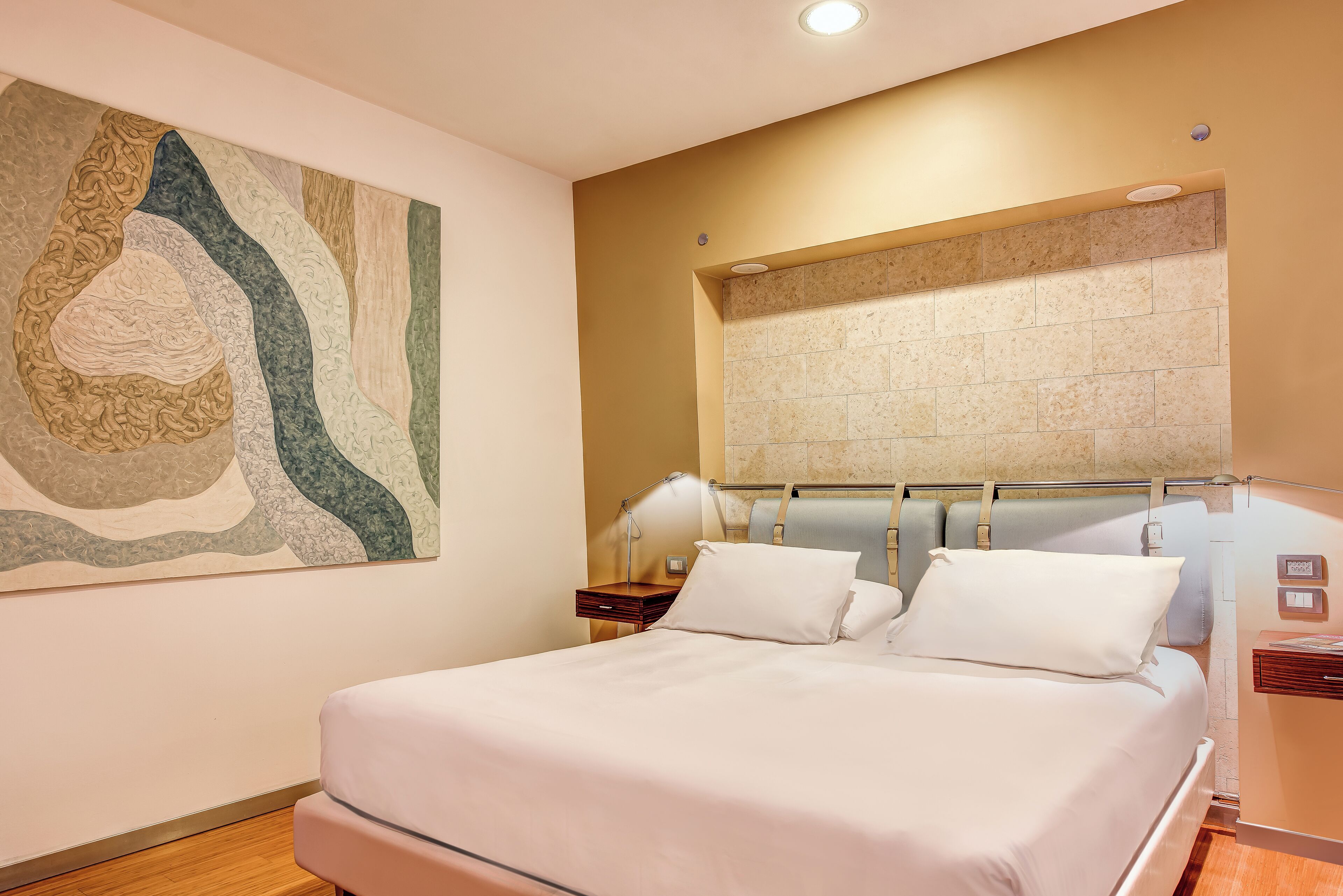 Cama bem arrumada num quarto de hotel, com um quadro abstrato grande na parede, cabeceira com prateleira embutida e mesa de cabeceira com abajur.