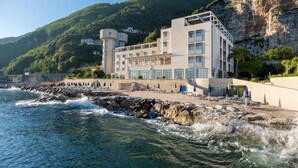 A coastal hotel with a rocky shoreline, a pool area with lounge chairs, and a mountain backdrop. - Towers Hotel Stabiae Sorrento Coast (Castellammare di Stabia)