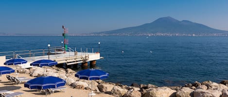 A beach with lounge chairs and umbrellas, a pier with a red and green structure, and a mountain in the background.