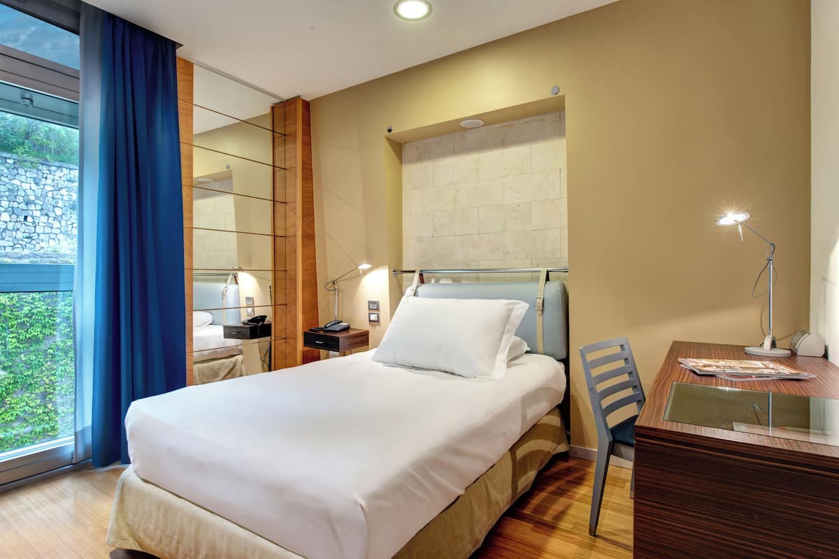 Ein Hotelzimmer mit einem Bett, einem Schreibtisch, einem Stuhl, einem Nachttisch, einer Lampe und einem Vorhangfenster.