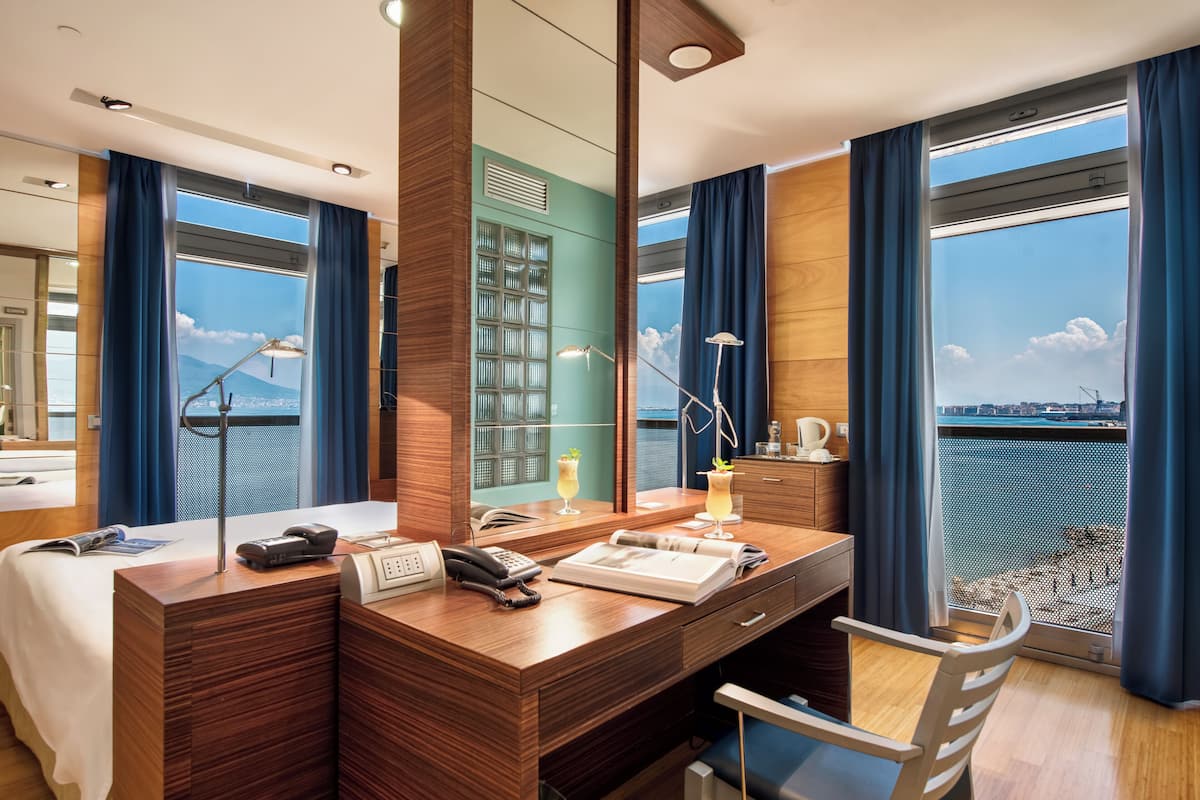 Ein Hotelzimmer mit Schreibtisch aus Holz, Telefon, Laptop, Stuhl, Minibar und einem großen Fenster mit Meerblick.