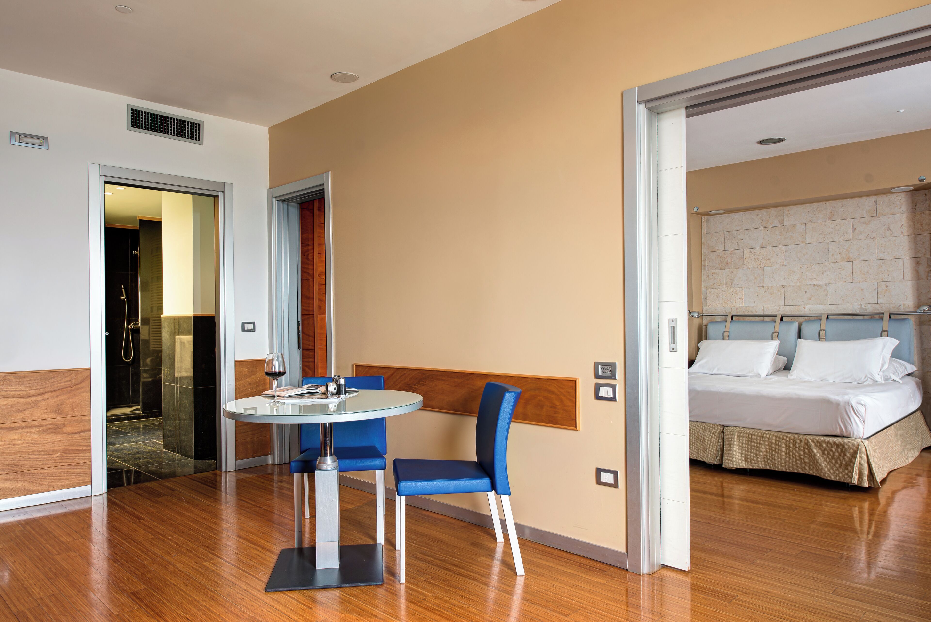 Une chambre d’hôtel avec un lit, une table ronde entourée de chaises, un plancher en bois et une salle de bain visible à travers une porte ouverte.