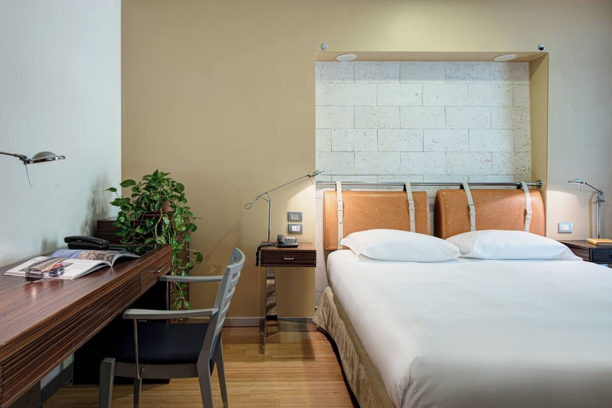 Ein Hotelzimmer mit einem Holztisch, einem Sessel, einem Bett mit weißer Bettwäsche, einem Nachttisch und einer Topfpflanze.