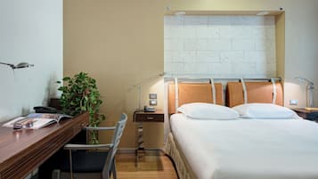Ein Hotelzimmer mit einem Holztisch, einem Sessel, einem Bett mit weißer Bettwäsche, einem Nachttisch und einer Topfpflanze.