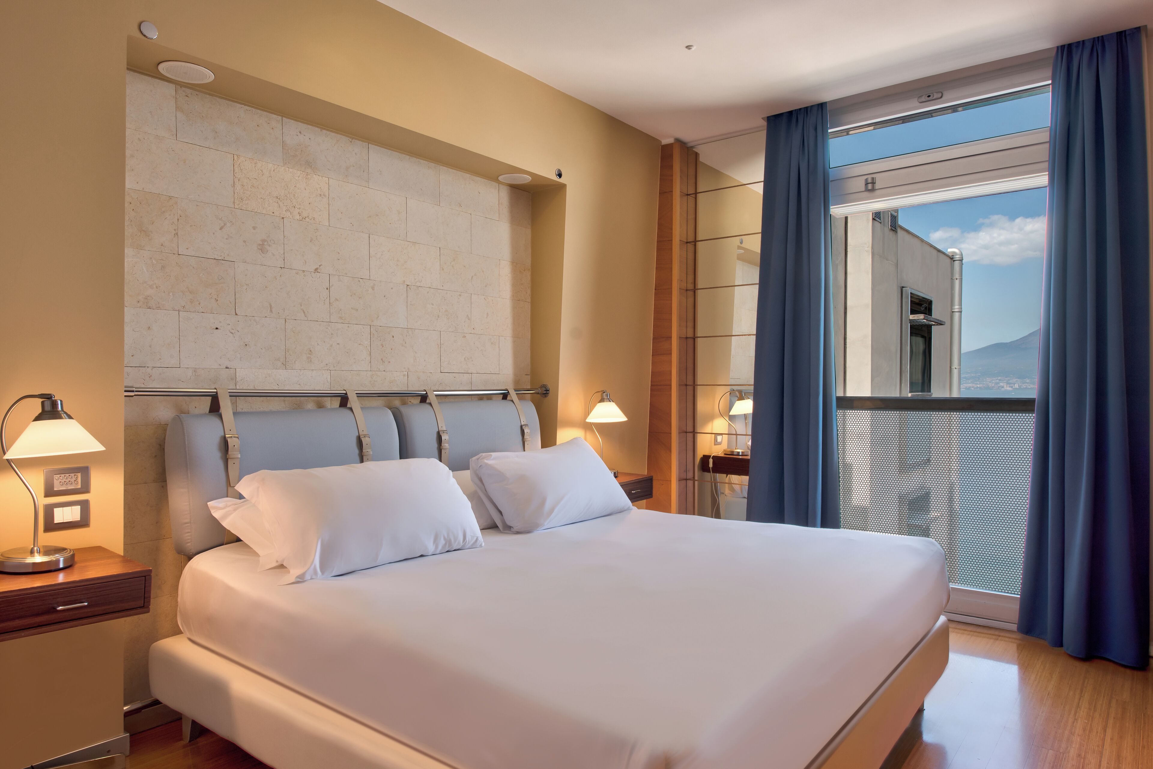 A hotel room with a large bed, a headboard, a nightstand with a lamp, and a view of the city through a balcony with blue curtains.