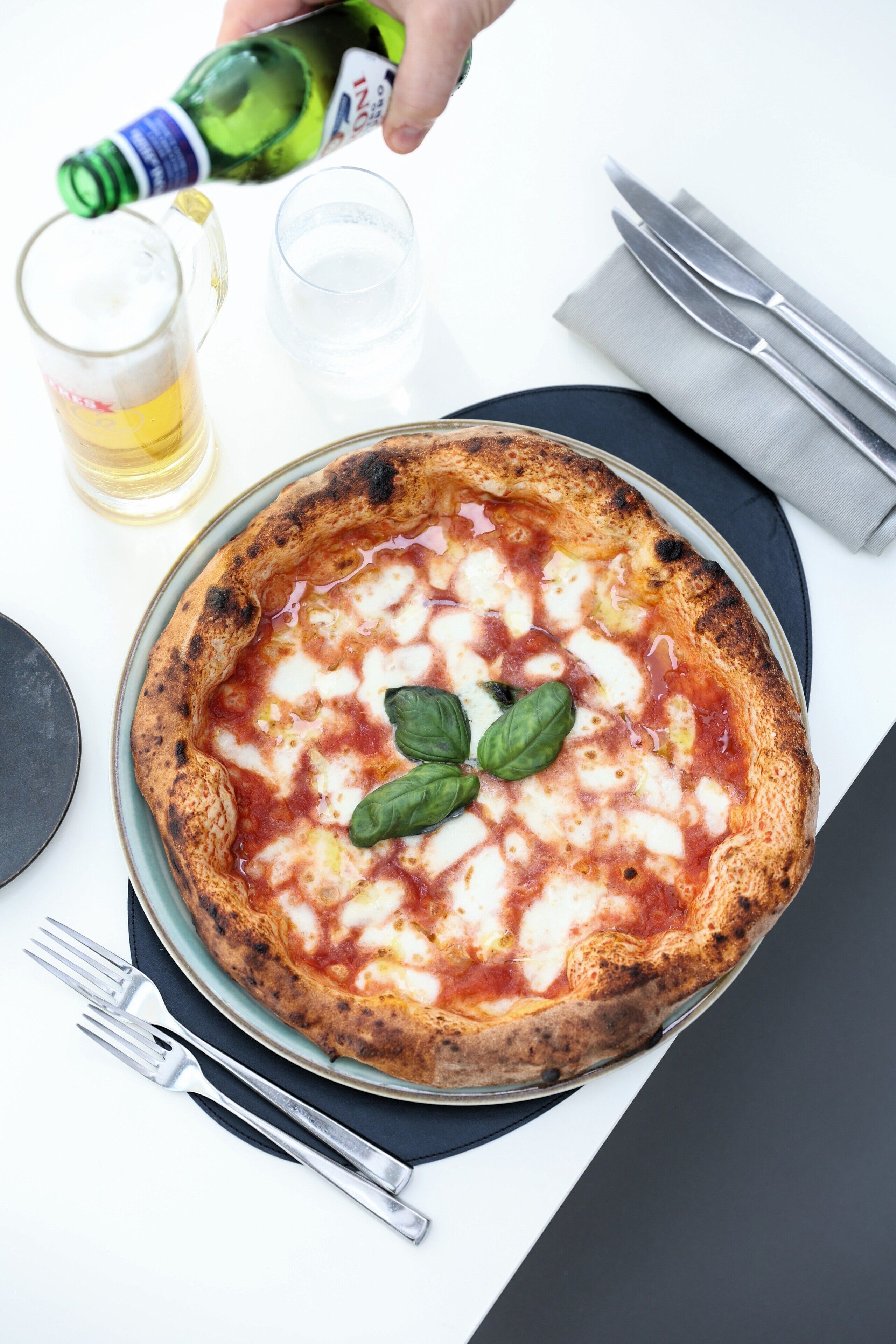Une pizza garnie d’une feuille de basilic, posée sur une assiette, avec une serviette et des couverts à côté.