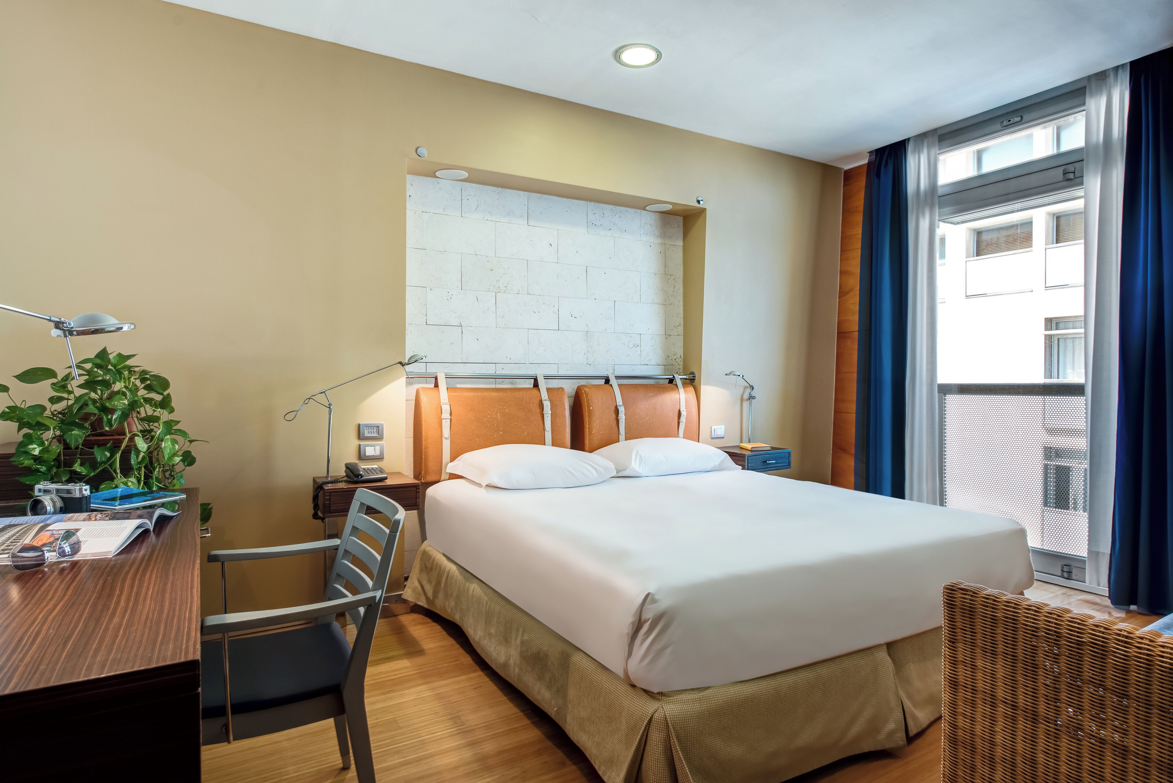 Hotelkamer met een groot bed, een bureau met stoel, een plant, een raam met gordijnen en een muur met baksteenmotief.