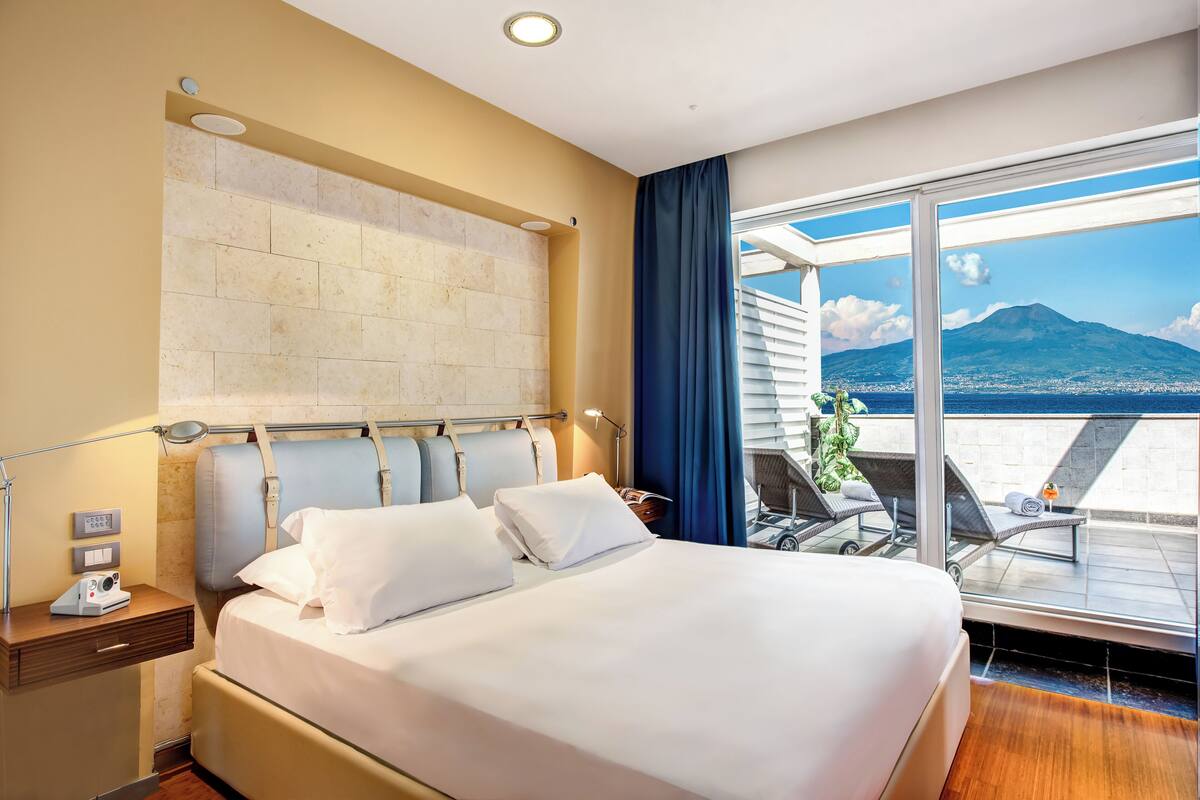 Ein Hotelzimmer mit einem großen Bett, einem Nachttisch, einem Schreibtisch mit Lampe sowie einem Balkon mit Blick auf Stadt und Berge.