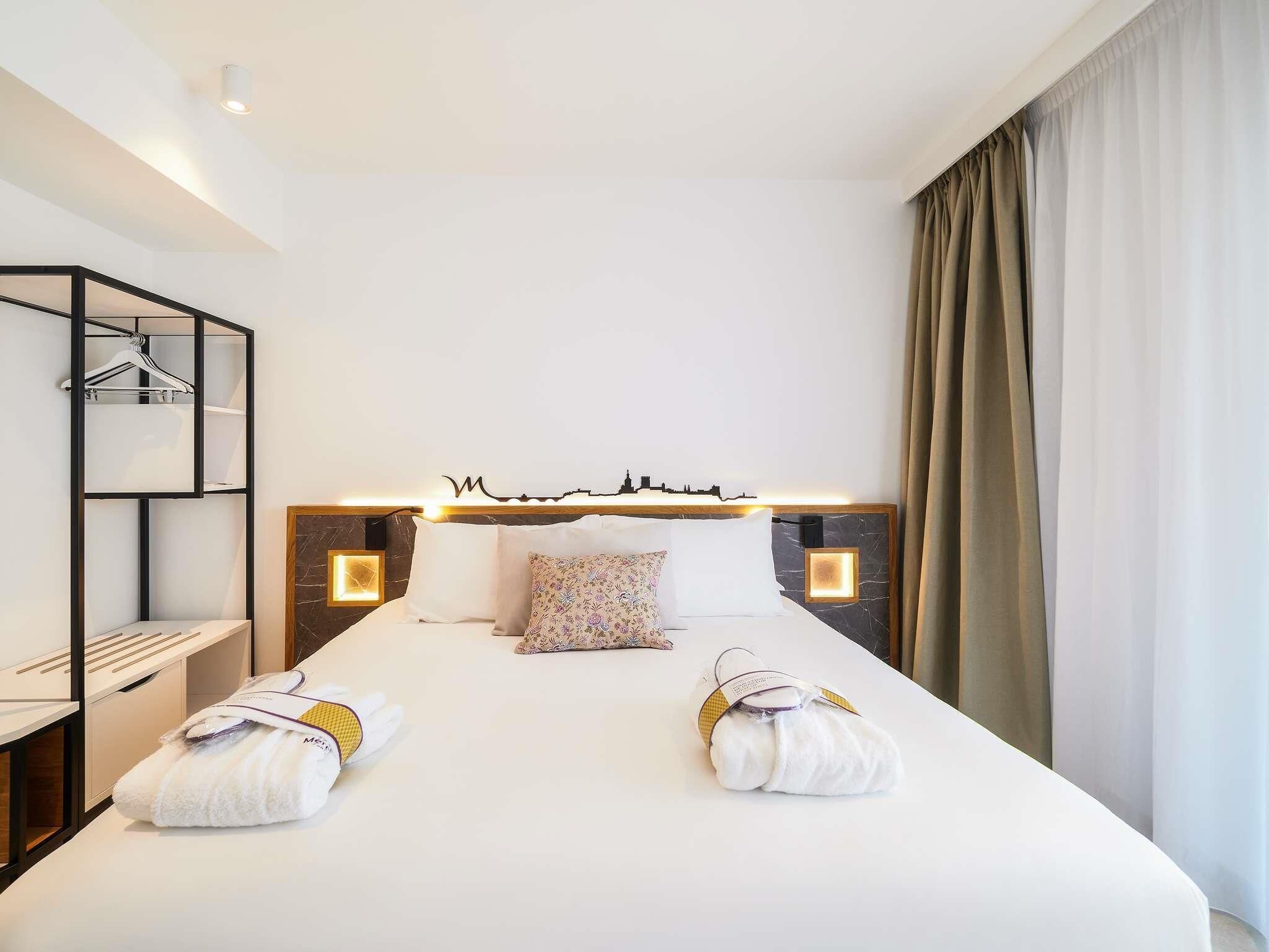 A hotel room with a large bed, two bedside tables, a headboard with a city skyline design, and a wardrobe with a hanging rack.