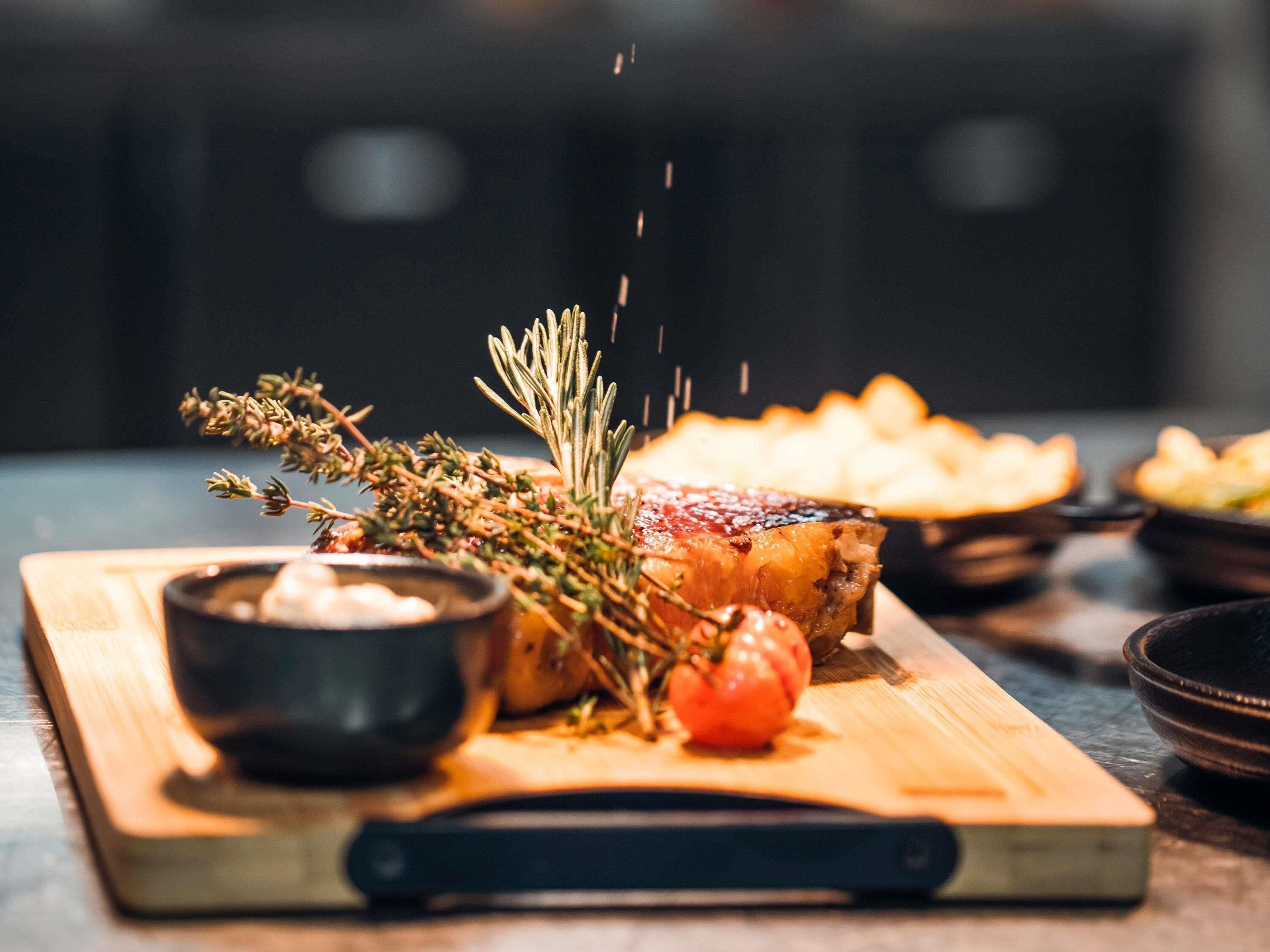 A wooden cutting board with a roasted meat, herbs, and a small bowl of sauce.