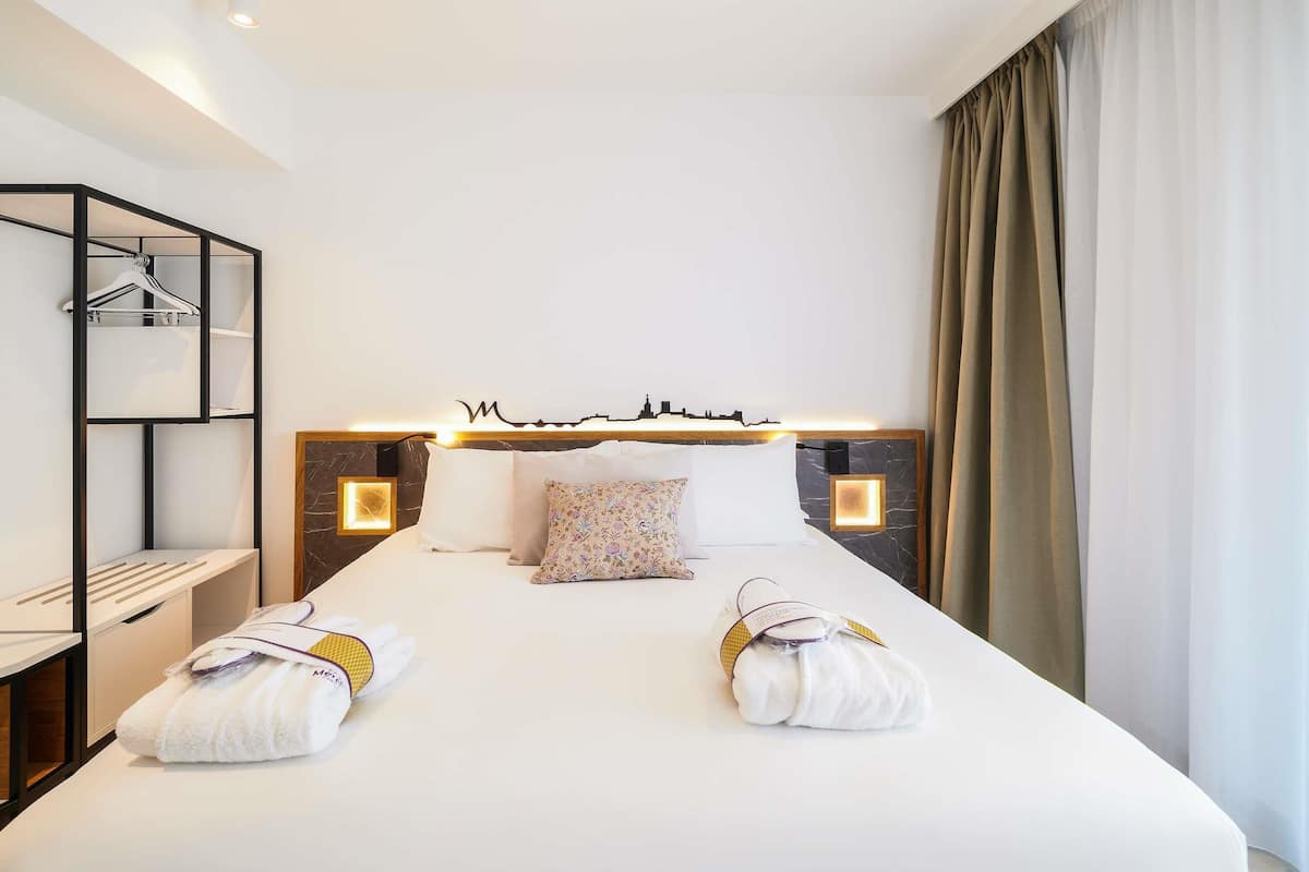 Ein Hotelzimmer mit einem ordentlich bezogenen Bett, einem Kopfteil mit Stadtblick-Motiv, Nachttischlampen, einem Kleiderschrank mit Kleiderstange und einem großen Fenster mit Vorhängen.
