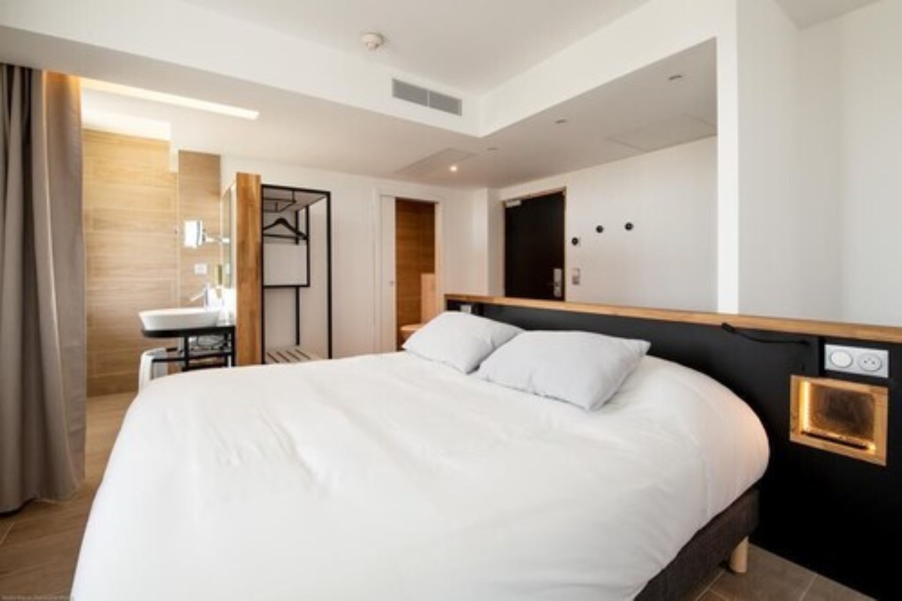 Una habitación de hotel moderna con una cama grande, una mesita de noche, un espejo y una zona de baño pequeña.
