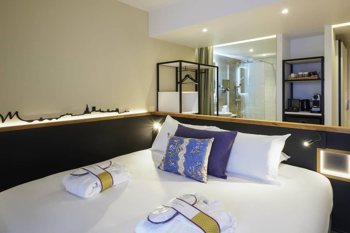 Ein Hotelzimmer mit einem großen Bett, einem Spiegel, einem sichtbaren Badezimmer durch eine offene Tür und einem Regal mit Toilettenartikeln.