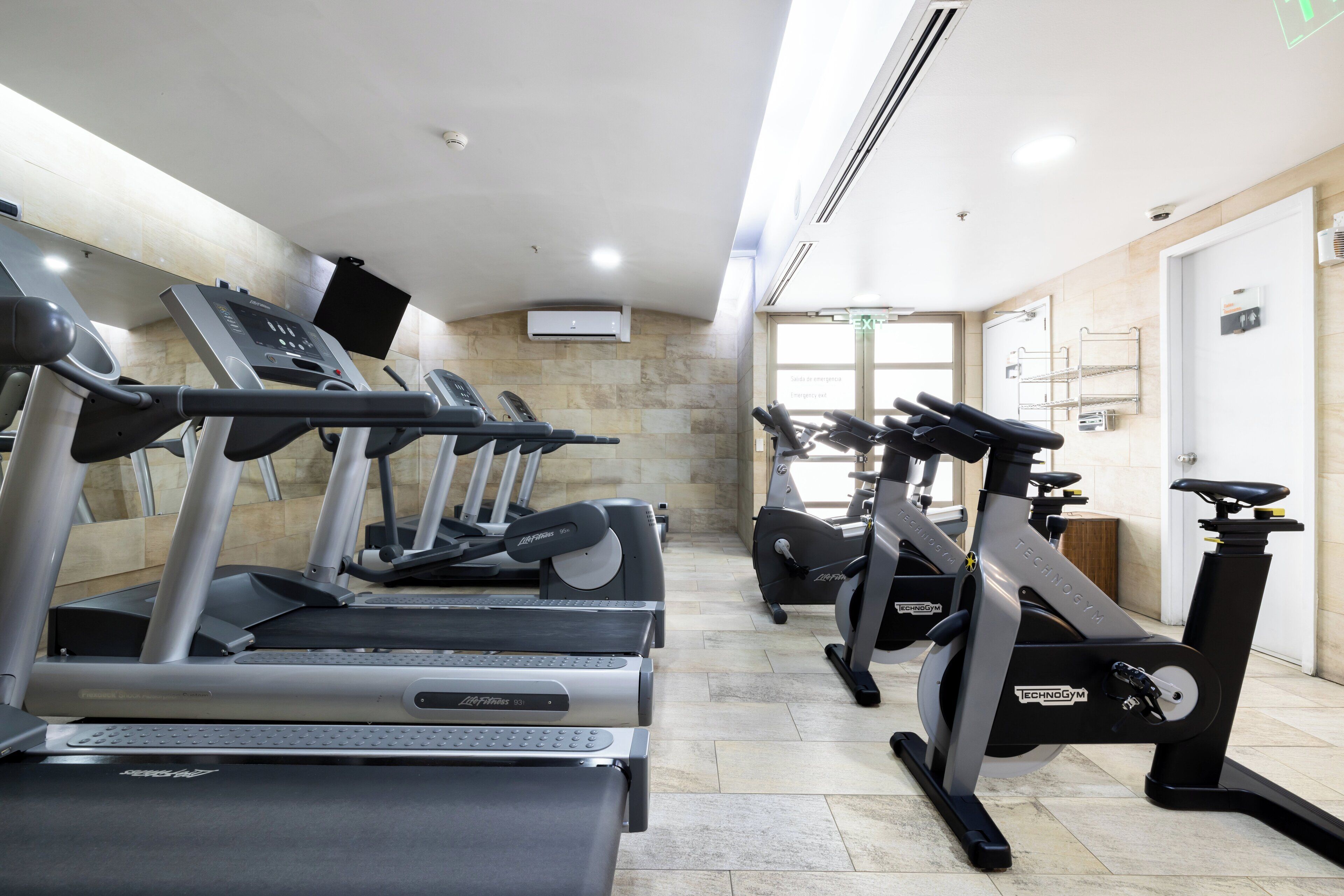 Un gimnasio con varias cintas de correr y bicicletas estáticas, suelo de baldosas y una puerta con letrero de salida.