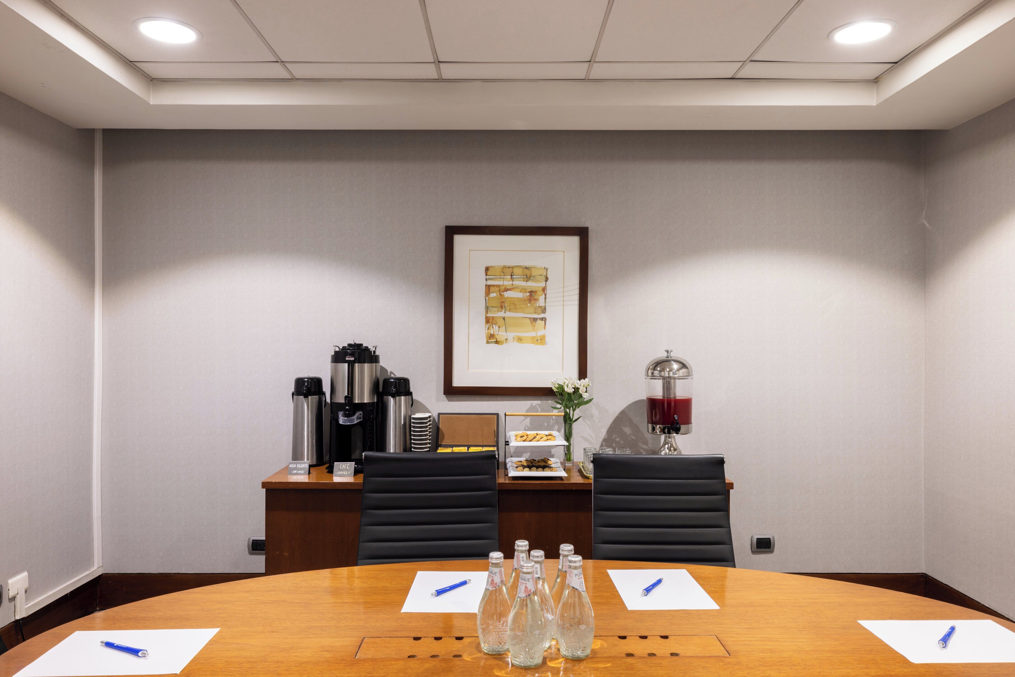 A conference room with a wooden table, black chairs, a coffee station, and a framed artwork on the wall.